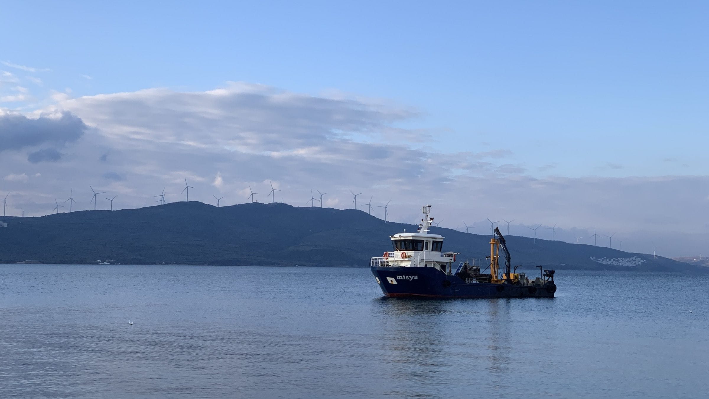 Misya Seafood’s blue work vessel, with a crane on its deck, on calm water in a bay, with wind turbines along the hills behind.